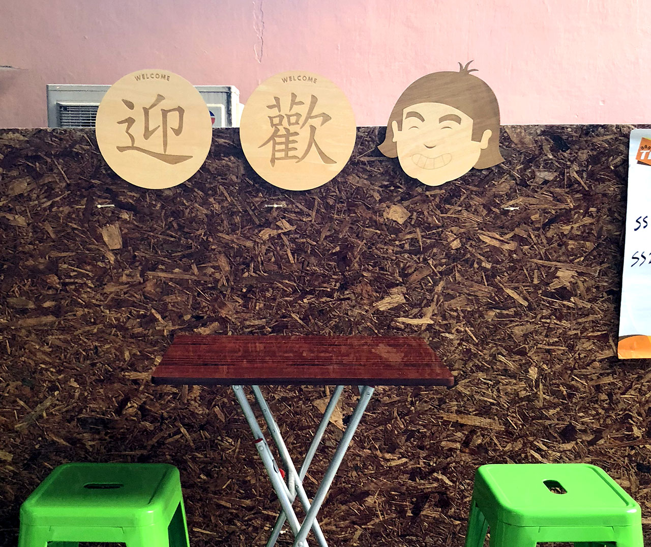 Outdoor seating showig two green stools, a foldable table, and above it, three lasercut wooden signs that say welcome in Chinese and feature a smiling person's head