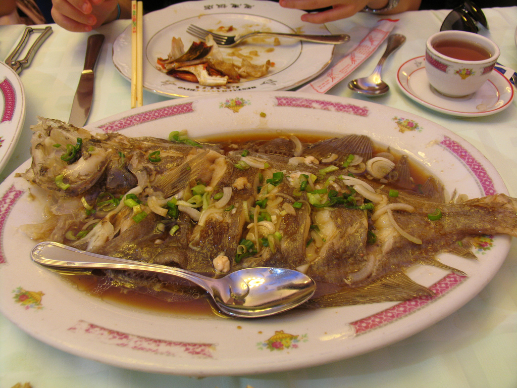 A steamed fish on a plate with ornate Chinese decorations while a hand hovers over a messy plate in the background